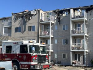 Fire-damaged apartment building with fire truck in front Front view of a multi-storey apartment building severely damaged by fire, showing extensive charring and structural collapse on the upper roof and balconies. Burnt debris hangs from the damaged sections, with broken windows and blackened siding. A Grande Prairie Fire Department (GPFD) red ladder truck with "Proudly Serving Since 1915" markings is parked in front, lights off, with caution tape, potted plants, and debris scattered on the ground under a bright blue sky.