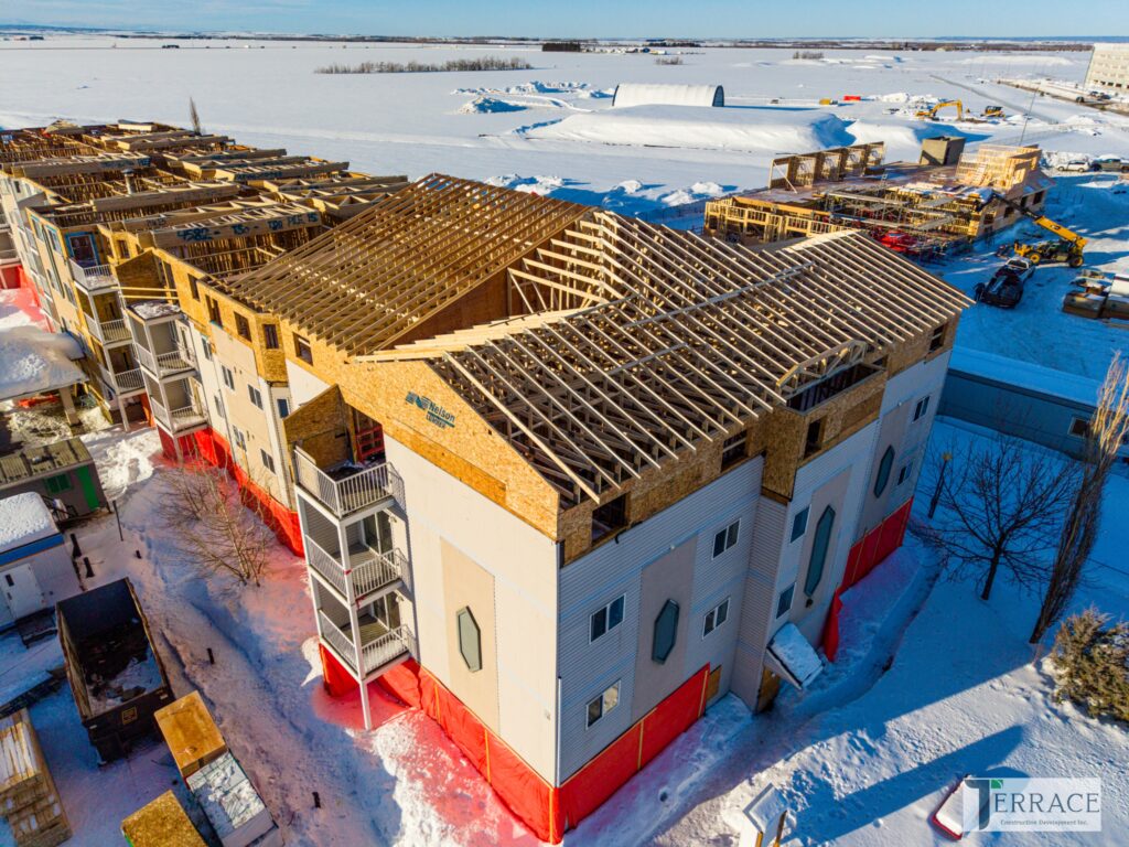 Aerial view of a large multi-family apartment building under construction in a snowy prairie landscape. The structure features a gabled roof with exposed wooden roof trusses, multi-level framed floors, balconies with railings, and red protective wrapping around the lower exterior. Surrounding area shows snow-covered ground, nearby industrial buildings, and distant cityscape under a clear blue sky