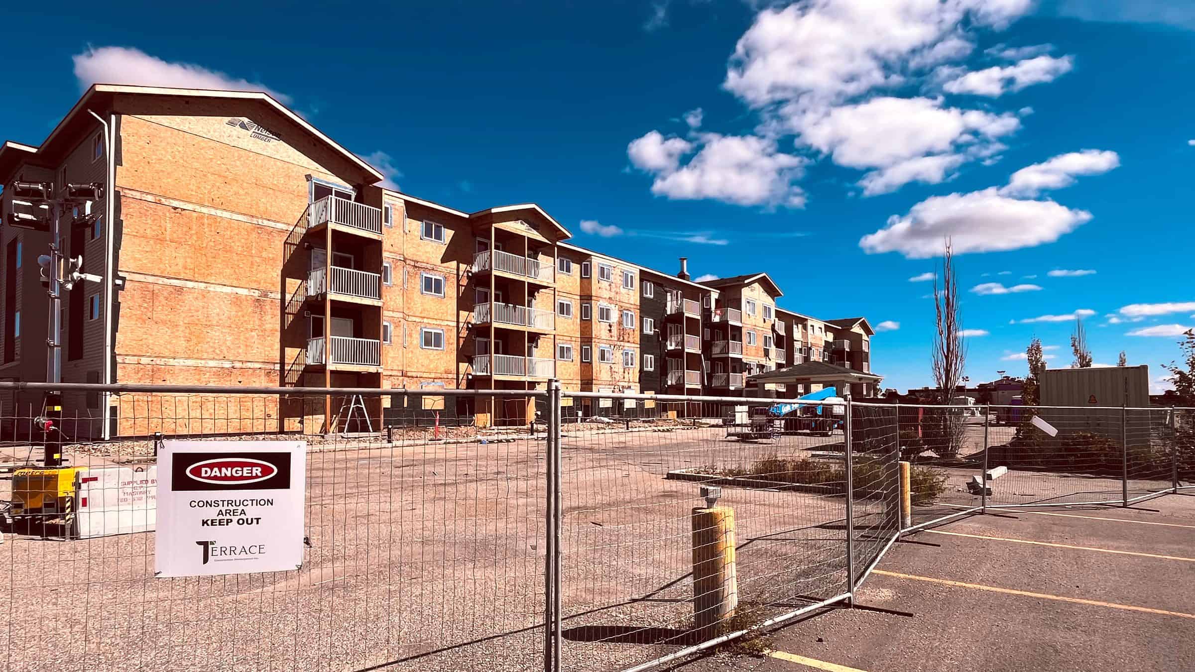 Margaret Edgson Manor in August 2026, stripped back to bare sheathing behind construction fencing, seen across the parking lot