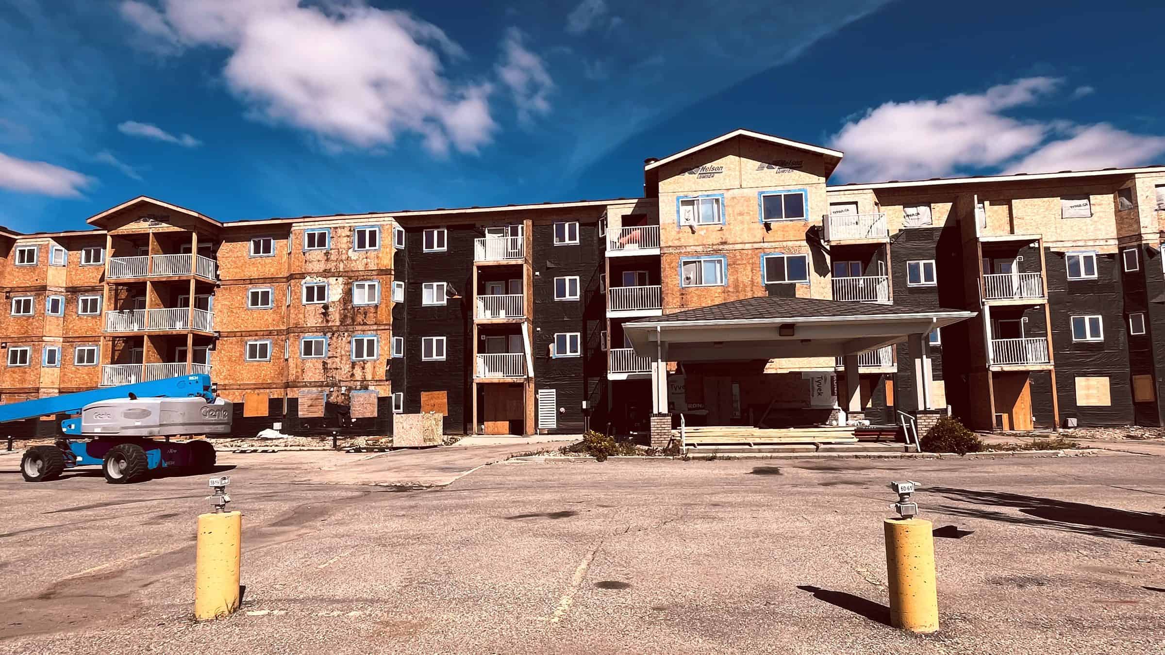 Margaret Edgson Manor in August 2026, its old siding stripped away to bare sheathing across all four storeys, with a boom lift parked out front