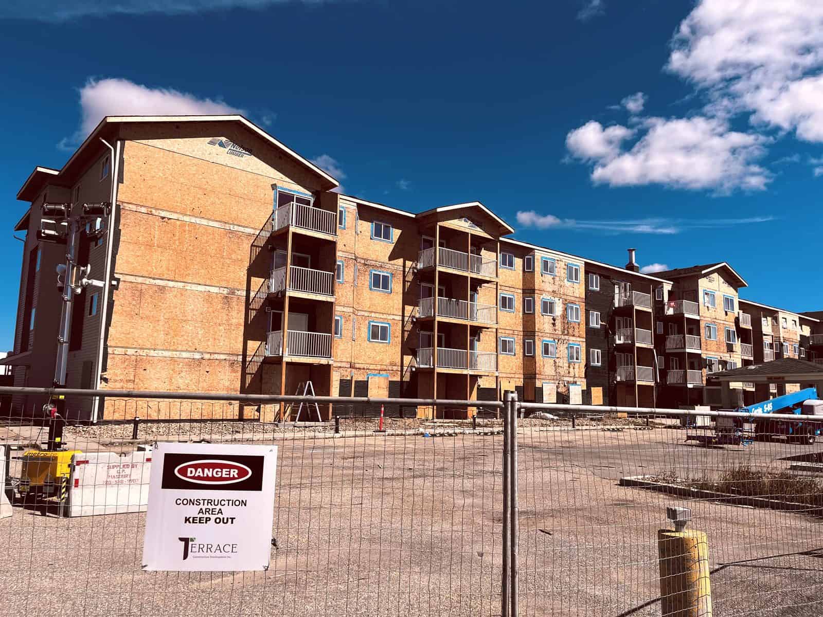 Margaret Edgson Manor from the southwest in August 2026, with the old siding stripped away to bare sheathing, balconies in place, behind construction fencing